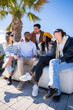 © CarlosBarquero - Vertical. Multicultural group of friends sit together on a bench outdoors in a bright residential district, chatting and enjoying leisure time under palm trees.