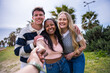 © CarlosBarquero - Three diverse young friends smile while holding the viewer's hand and leading them forward through a sunny green park, conveying friendship and shared adventure.