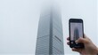 © joeycheung - Hand holds phone capturing foggy skyscraper, mist obscures its towering presence.