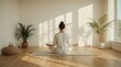 © Atif - Woman practices seated meditation on a mat facing a wall illuminated by soft morning sunlight between two potted palm plants.