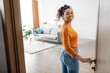 © Prostock-studio - Joyful Smiling Black Woman Opening Door With Happiness, Standing In Modern Living Room, Welcoming Guests, In Cozy Home Environment. Home Interior Concept.
