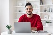 © Prostock-studio - Smiling Young Man Of Middle Eastern Descent Working On Laptop At Home Office, Sitting At Table With Notebook And Plant, Engaged In Remote Work, Promoting Business Concept.