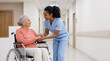 © Someone Cooked Here - young black female doctor holding hands with elderly white female patient seated in wheelchair in clinic hallway