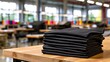 © Daria - Neatly folded stack of black fabric shirts resting on a wooden table inside a brightly lit garment manufacturing factory environment