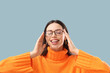 © Pixel-Shot - Headache relief and stress management, woman with migraine touching temples while wearing glasses and orange sweater on a blue background