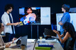 © ultramansk - A multiethnic group of professionals discuss the report with a remote leader on video in a dark blue lit conference room at night, an image of hybrid teamwork across distance