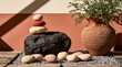 © David Giraud - Une composition minimaliste d'inspiration zen, associant des pierres et un pot en terre cuite avec plante verte contre un mur ensoleillé, vue en gros plan.