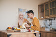 © Alfian Muzaki - Young Asian Muslim couple cooking together in a warm modern kitchen, smiling as they prepare a meal with eggs, fruit and fresh ingredients, sharing a relaxed, domestic and affectionate moment at home.