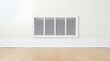 © Pixels Stock - White rectangular air vent grill installed on light beige wall with white baseboard and wooden floor in room interior.