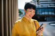 © Davor - Smiling woman using smartphone outside modern office building