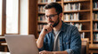 © thiago - A young man with glasses and a beard sits in a library, focused on his laptop, with bookshelves in the background.