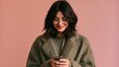 © Yassine - Smiling young woman with dark hair wearing green coat looks down while holding smartphone against plain solid pink studio background
