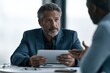 © Vilaysack - A senior executive holds a tablet and listens attentively while a colleague gestures during a formal office meeting.
