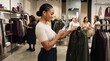 © mashimara - Chic Shopping Experience: A stylish woman engrossed in her smartphone browses a curated selection of fashionable apparel in a modern retail boutique.