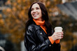 © deagreez - Smiling woman in leather jacket holding a takeaway coffee cup on an autumn city walk, cozy orange sweater and relaxed happy expression