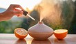 © Maek - hand holding dropper with organic sweet orange essential oil above ceramic diffuser mist spraying in sunlight wellness and relaxation concept fresh and uplifting mood