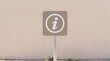 © amera - Beige square information sign featuring a white information symbol inside a circle mounted on a metal post against a beige wall.
