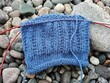 © Elena - Blue knitted fabric on circular needles resting on smooth pebbles. Handmade craft and knitting hobby concept