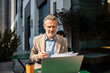 © Davor - Mature businessman reviewing documents on laptop at outdoor cafe