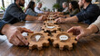 © Christophe - Diverse colleagues align a long chain of wooden gears with lightbulb and communication icons across a shared table, illustrating teamwork, collaboration, connection, ideas and coordinated business wor