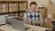 © Krakenimages.com - Man eating salad with fork while seated at laptop among stacked cardboard boxes and parcel shelves in a building; concentration.