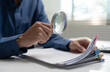 © Father_Studio - A professional male businessman uses a magnifying glass to carefully inspect a thick stack of paperwork. The auditor reviews financial documents and business contracts at his desk.