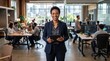 © Pravinrus - A smiling African American female executive in a business suit holding a digital tablet while standing in a busy, modern open-plan tech office with coworkers working in the background.