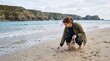 © Ednardo - Coastal Collection: A woman with tousled hair searches the sandy shore for shells, her casual attire suggesting a relaxed day by the sea, with rugged cliffs framing the tranquil blue ocean.