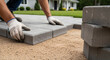 © jendralbiribiri - Paver installation worker laying gray concrete bricks on a sandy surface for a residential patio landscaping project in summer
