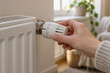 © AiHRG Design - Close-up of a hand turning a white radiator thermostat valve in a bright, cozy home interior with soft natural light.