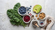 © Sasi - Flat lay top view of healthy superfoods assortment including blueberries, sliced avocado, pomegranate seeds, kale, chia seeds, and walnuts on grey stone background