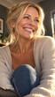 © Adrianna - A smiling middle aged blonde woman looks aside happily in a sunlit indoor setting, wearing a beige knit cardigan, white top, and blue jeans