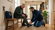 © Maryia - Adult son helping father choose comfortable walking shoes before a walk, showing mobility support, family care, active aging, trust and practical independence at home together.