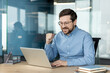 © Tetiana - Smiling businessman celebrating success while working on laptop in modern office with confident gesture, glass wall background, desk workspace, and professional business atmosphere