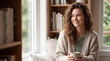 © Aneela - Smiling woman with curly hair enjoying a warm drink at home, sitting by a bright window in a cozy reading nook with bookshelves and soft natural light indoors