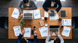 © Lukman - Overhead flat lay of a diverse business team collaborating around a wooden meeting table with laptops, digital tablets, and financial chart reports during a corporate strategy session