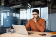 © Liubomir - Man in orange shirt sits at desk with laptop in modern glass office, resting hand on chin while concentrating on screen, with man, office, laptop, desk detail, showing business, worker, professional