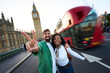 © CarlosBarquero - Happy interracial couple posing on Westminster Bridge with Big Ben and a moving red double-decker bus behind. Wide-angle travel portrait conveying excitement, friendship and London energy.