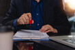 © khunkornStudio - Business person stamping approval on document at office desk with laptop and clipboard for compliance verification and administrative workflow