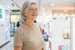© luciano - Smiling mature adult woman with gray hair wearing glasses shopping for cosmetics in a modern department store, enjoying a leisure lifestyle activity