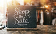 © velikiyzayats - Chalkboard sign with text Shop the Sale on wooden table in clothing boutique store retail promotion clearance discount offer merchandising announcement commerce