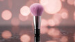 © xinxia - A soft pink makeup brush with metallic ferrule rests on a reflective dark surface, against a warm pink bokeh background.