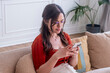 © simona - Young woman sitting on a sofa in a bright living room, relaxed and focused while using her smartphone to text, browse or shop online, casual style, glasses and cozy home.