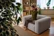 © djile - Woman stretching arms overhead and yawning while sitting comfortably on an armchair in a modern living room, embracing a tranquil morning routine at home