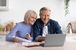 © Prostock-studio - Smiling Elderly Caucasian Couple Sitting Together At Kitchen Table, Engaged In Financial Planning While Using Laptop And Reviewing Papers, Enjoying Quality Time And Collaboration At Home.