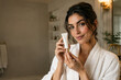 © new - Smiling young woman in bathrobe holding white cosmetic cream tube in modern bathroom