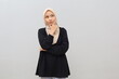 © ArvStd - A young Asian Muslim woman wearing a headscarf has a thoughtful, serious expression; she looks upward into the empty space above her, touching her chin while smiling as she imagines something.