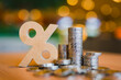 © sommart - Interest rate and compound growth concept with wooden percentage sign beside stack of money coins on warm bokeh background, representing bank deposit dividend, financial savings, and tax return.