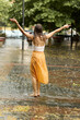 © Oleksandr - Back view of a carefree woman dancing barefoot on a wet paved sidewalk during heavy summer rain with hands wide open, feeling joy, freedom, and emotional release outdoors.