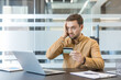 © Liubomir - Financial stress and online payment issues, man holding credit card looking at laptop with frustration in office. Economic hardship and personal debt management.